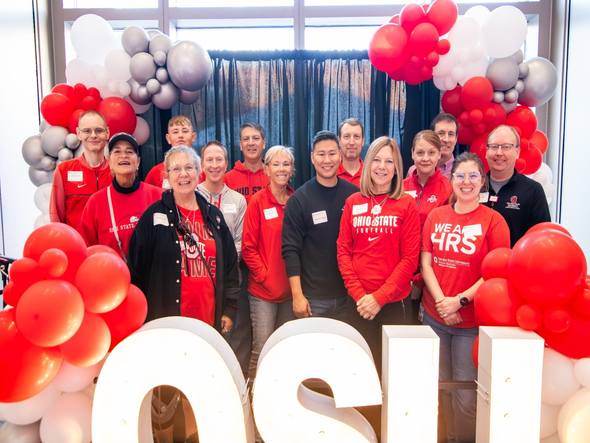 A group of people including HRS alumni in a group photo wearing Ohio State apparel. There are scarlet and gray balloons behind the people and the letters OSU in front.