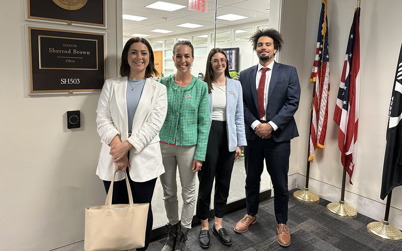 Three women and one man all dressed professionally in front of an office for U.S. Senator Sherrod Brown. There are flags in the background.