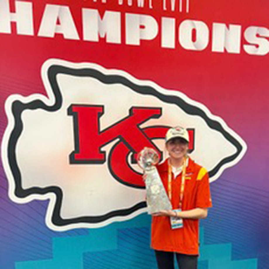 Kelly Weller wearing a red shit and hat while holding the Super Bowl trophy in from of a Kansas City Chiefs sign