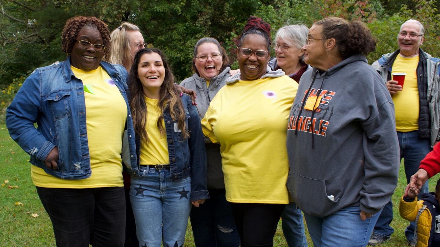 A group of eight joyful adults wearing casual attire, some in yellow shirts, laughing and chatting together in a park setting.