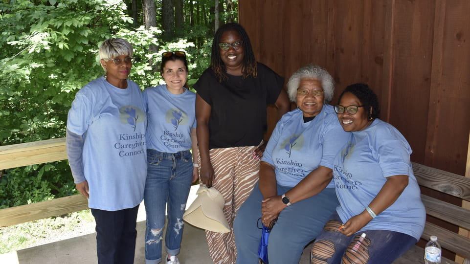 Five individuals wearing Kinship Caregivers Connect t-shirts sitting together on a bench, smiling in a wooded park setting.