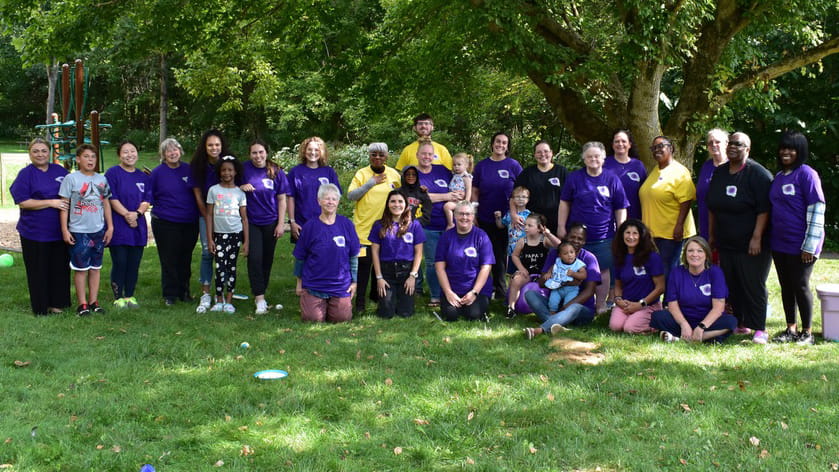 Group of diverse adults and children outdoors, wearing coordinating purple and white t-shirts, in a grassy park setting. Some individuals are standing, while others are kneeling or seated on the grass. They are grouped together, smiling broadly, at acommunity event. Trees and playground equipment are visible in the background.