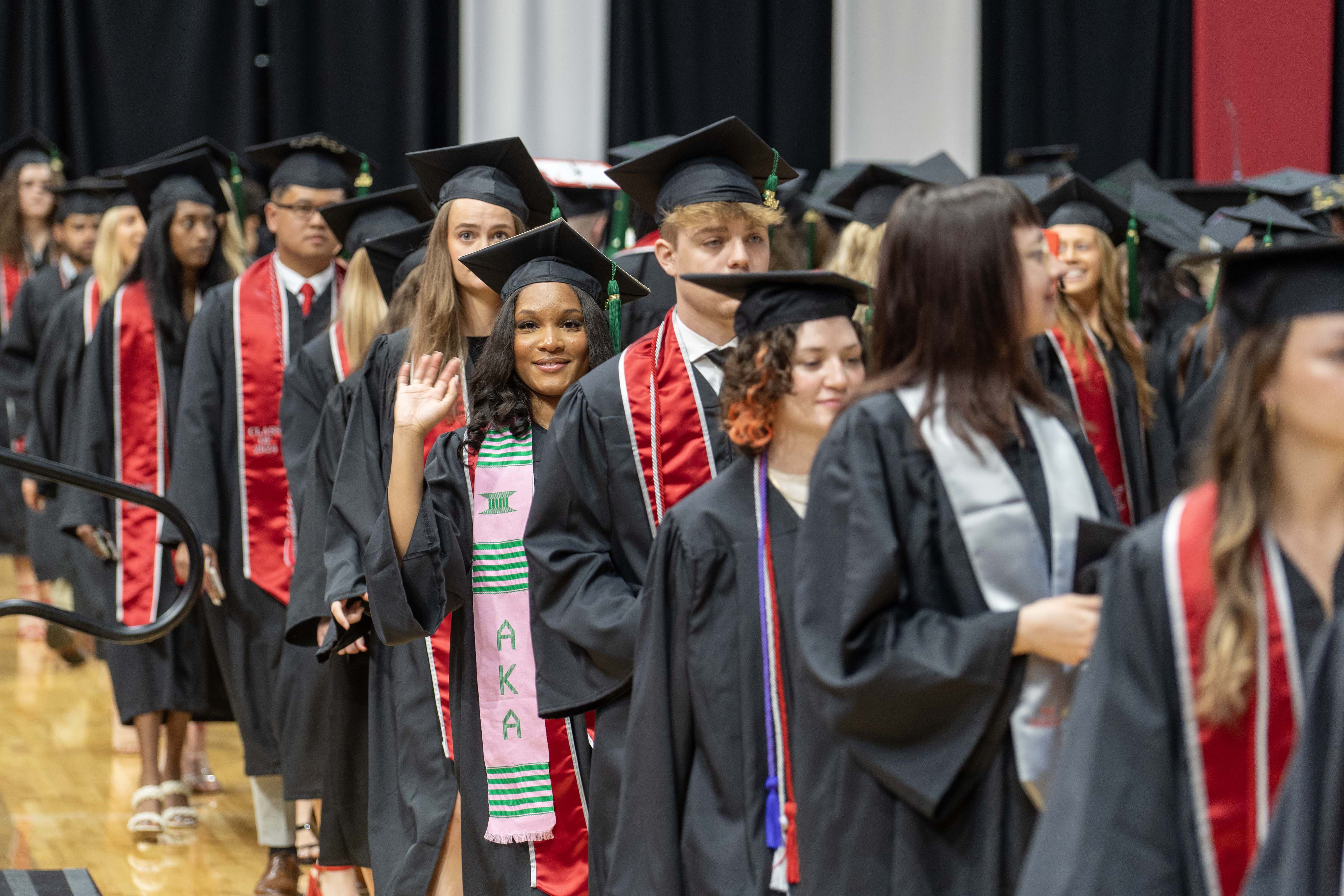 Graduates wearing caps and gowns walking in a procession during a graduation ceremony. One graduate is waving and smiling.