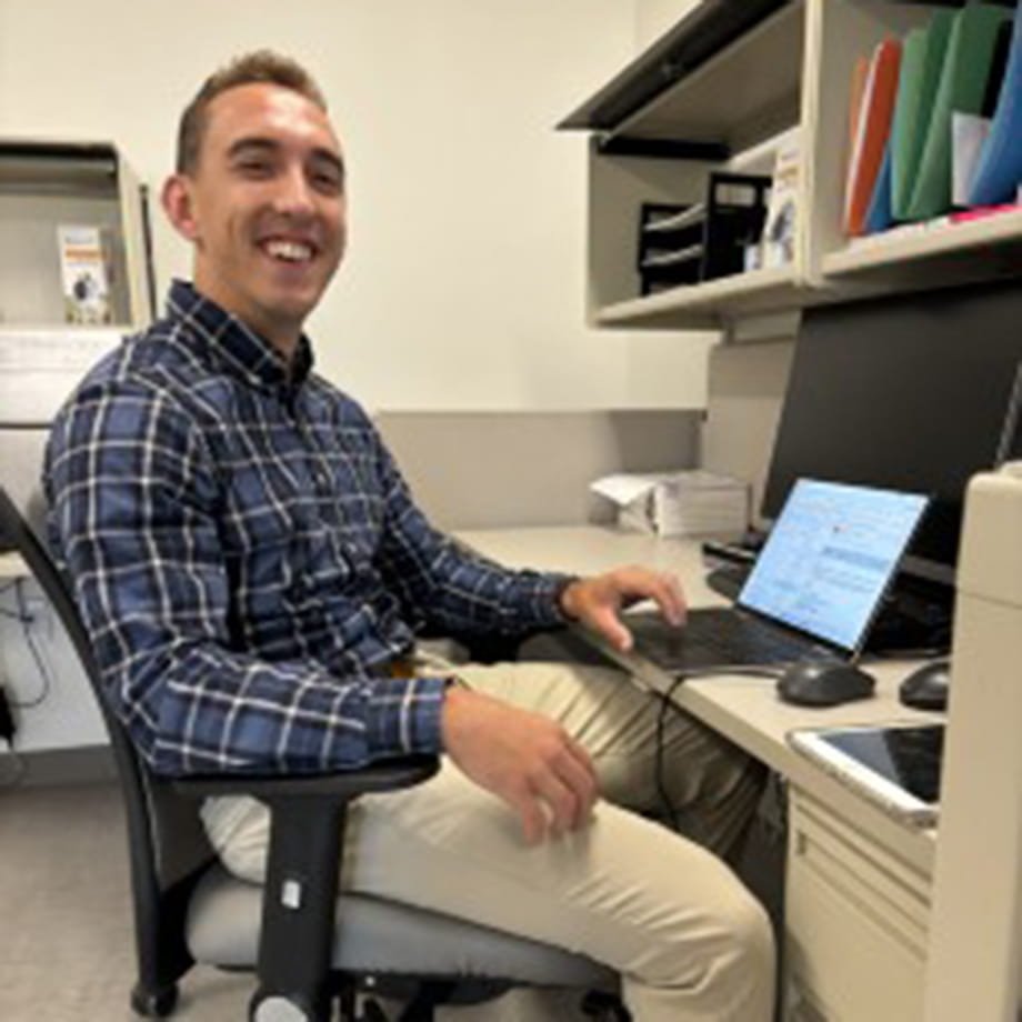 Dan Gilmore sitting at a desk with a computer, smiling at the camera in an office setting.