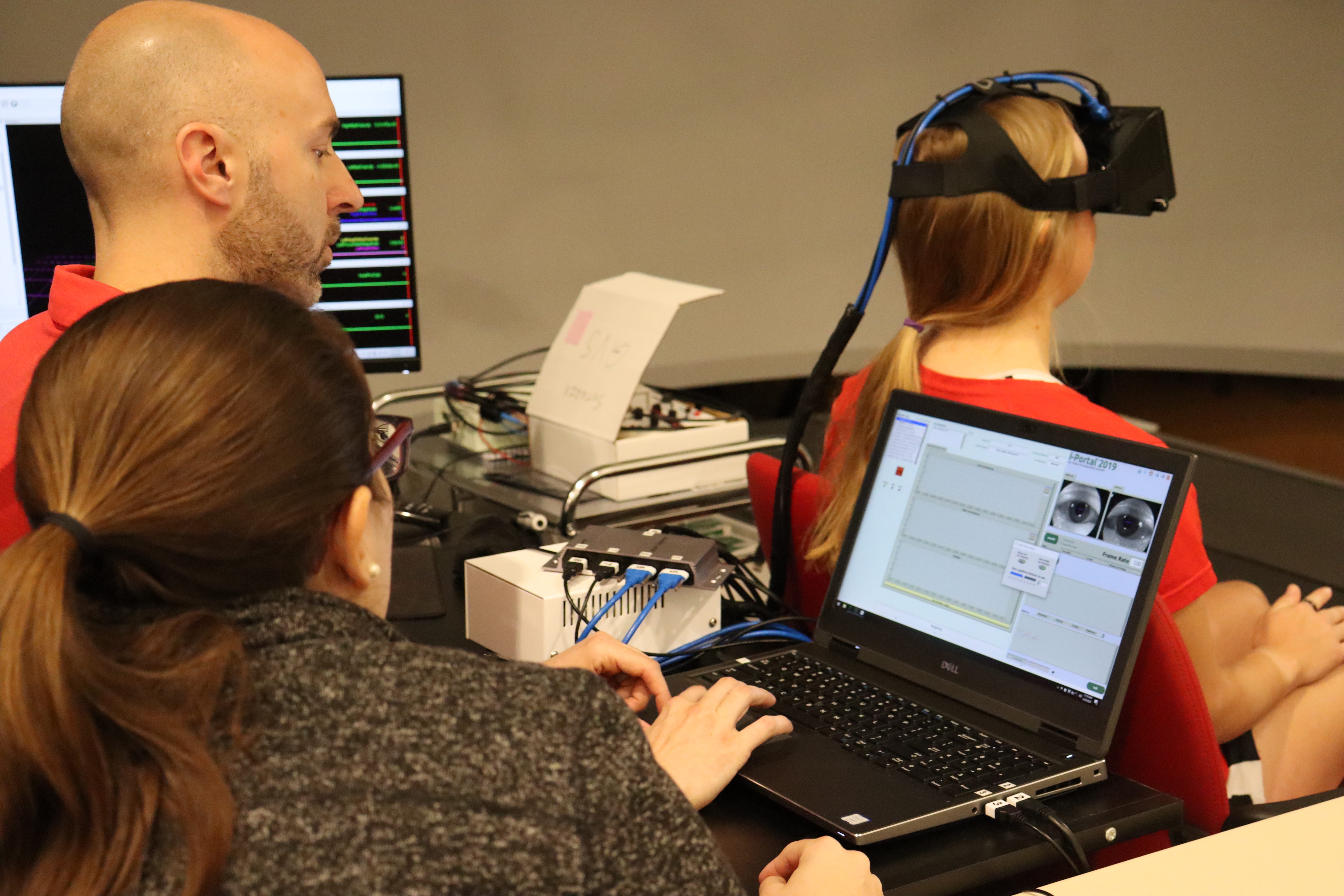 Two people are seated at a desk equipped with laptops and other electronic devices, engaged in a neuroscientific study. One participant is wearing a brain-computer interface headset. The screens display various graphs and data.