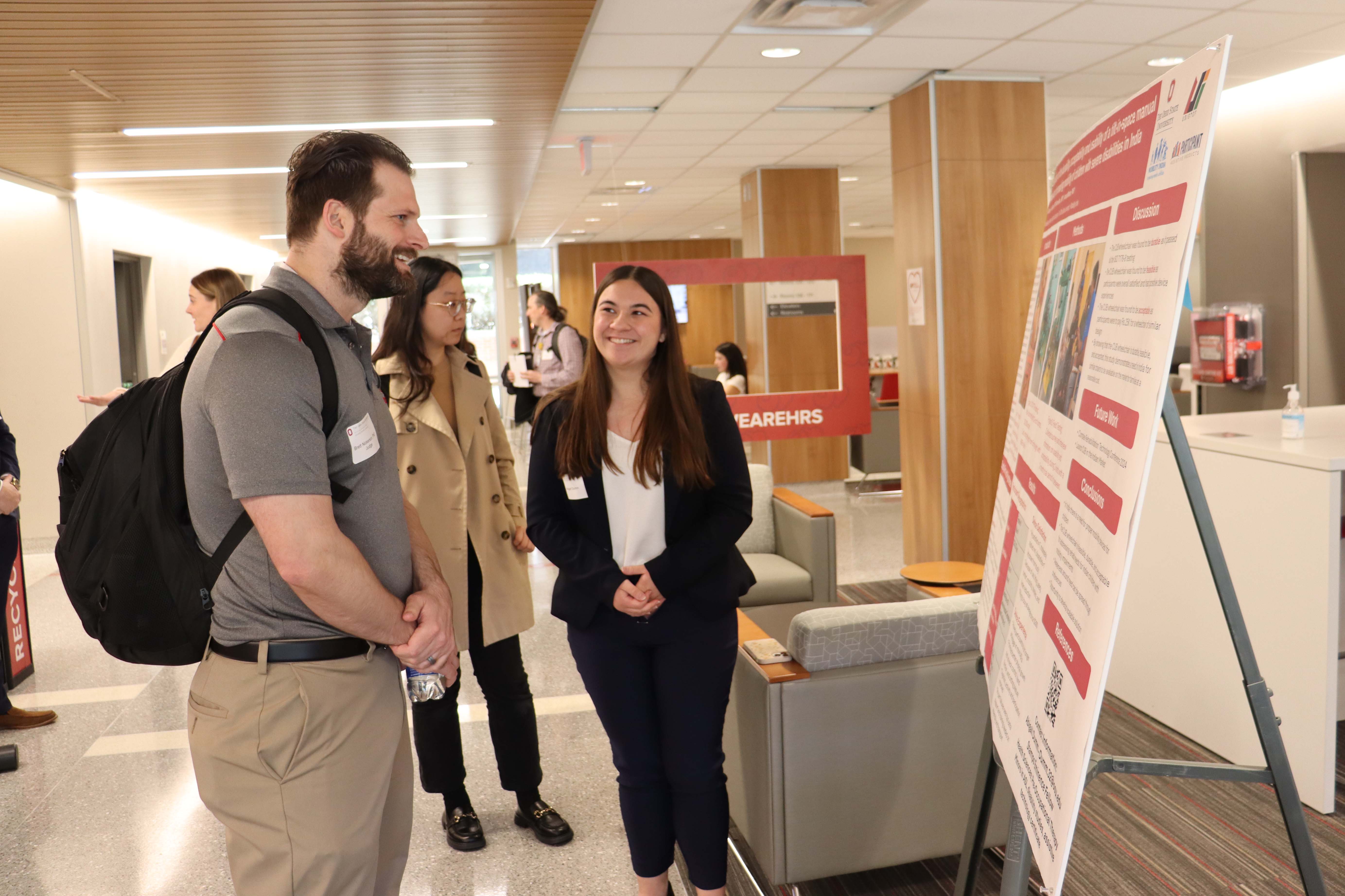 Two people conversing near a poster presentation in an indoor setting. One person stands next to a poster board explaining its content, while others, carrying backpacks, listen attentively at a research day presentation.
