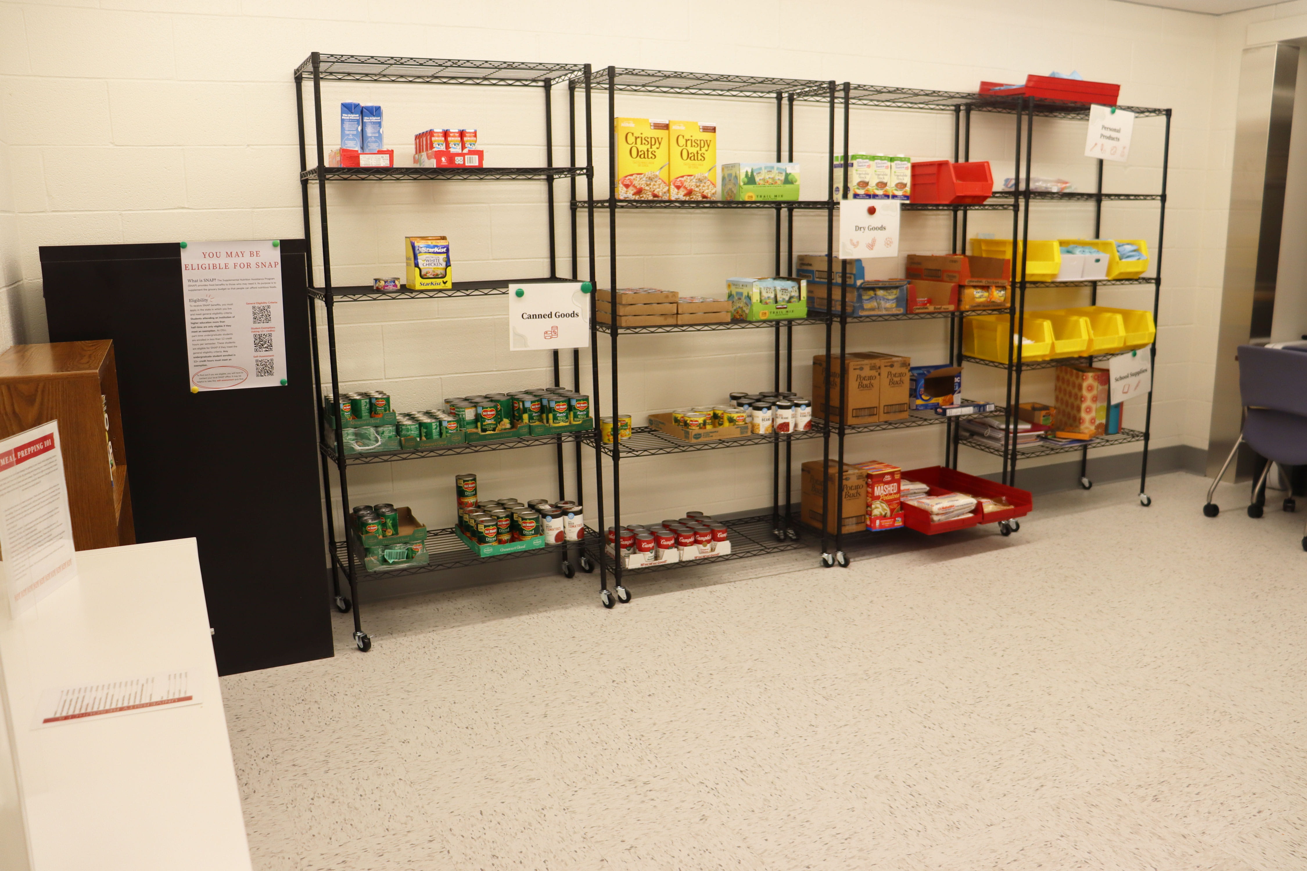 The image shows a food pantry with organized shelves stocked with various food items such as cereal boxes, canned goods, and snacks in a clean, well-lit indoor setting.
