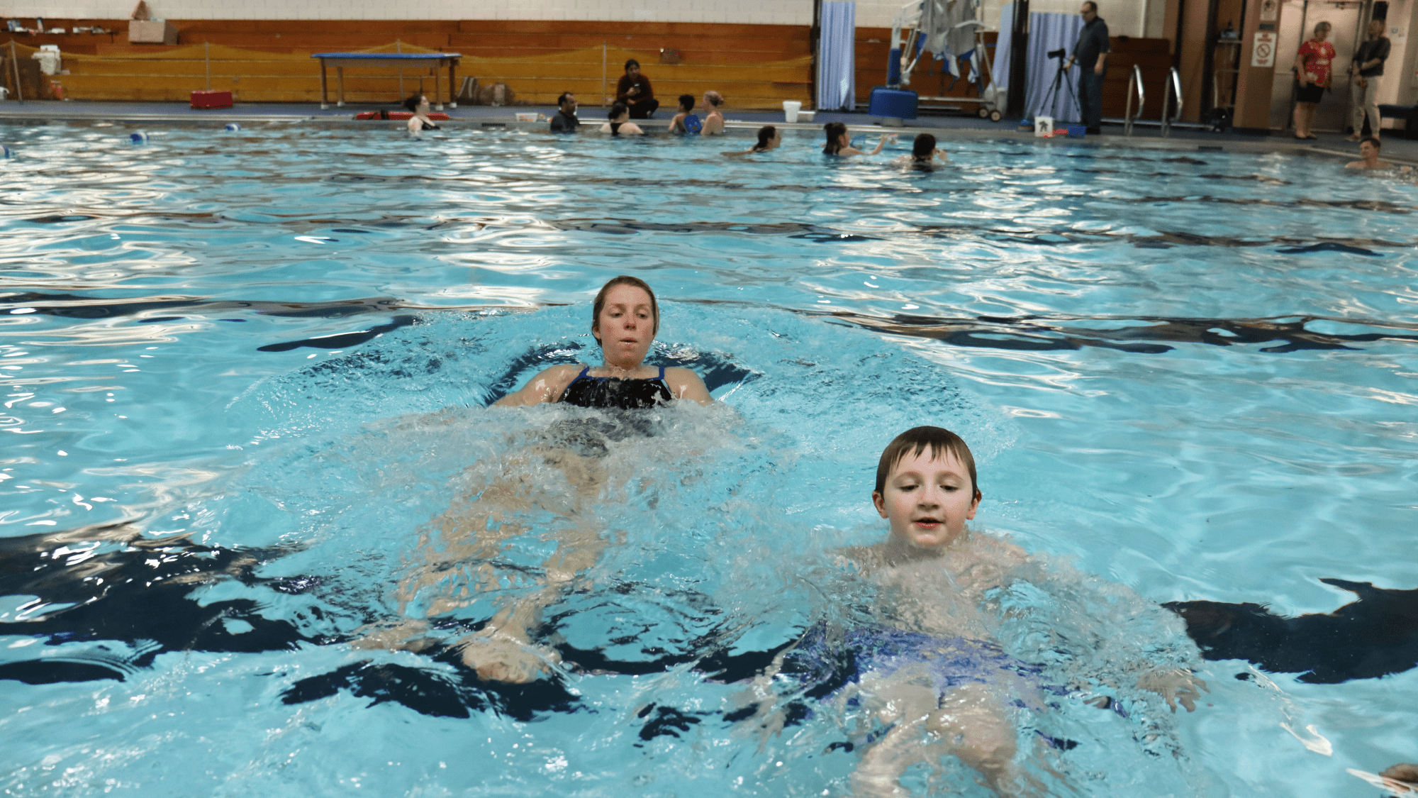 A child and swim instructor swimming backwards in a pool. There are other people in the background on the other side of the pool.