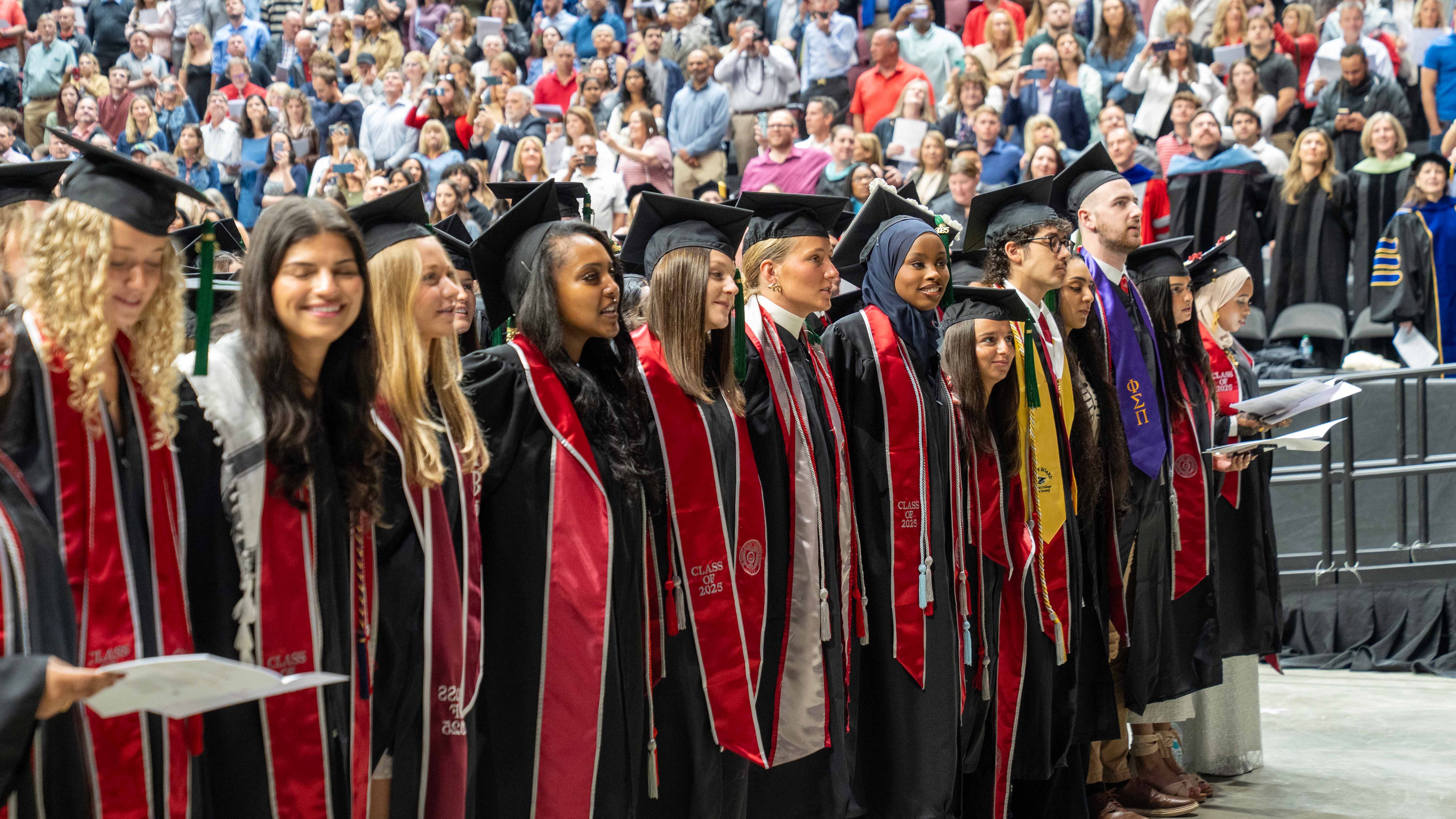 Graduates in caps and gowns standing during a ceremony, with a crowd of spectators in the background.