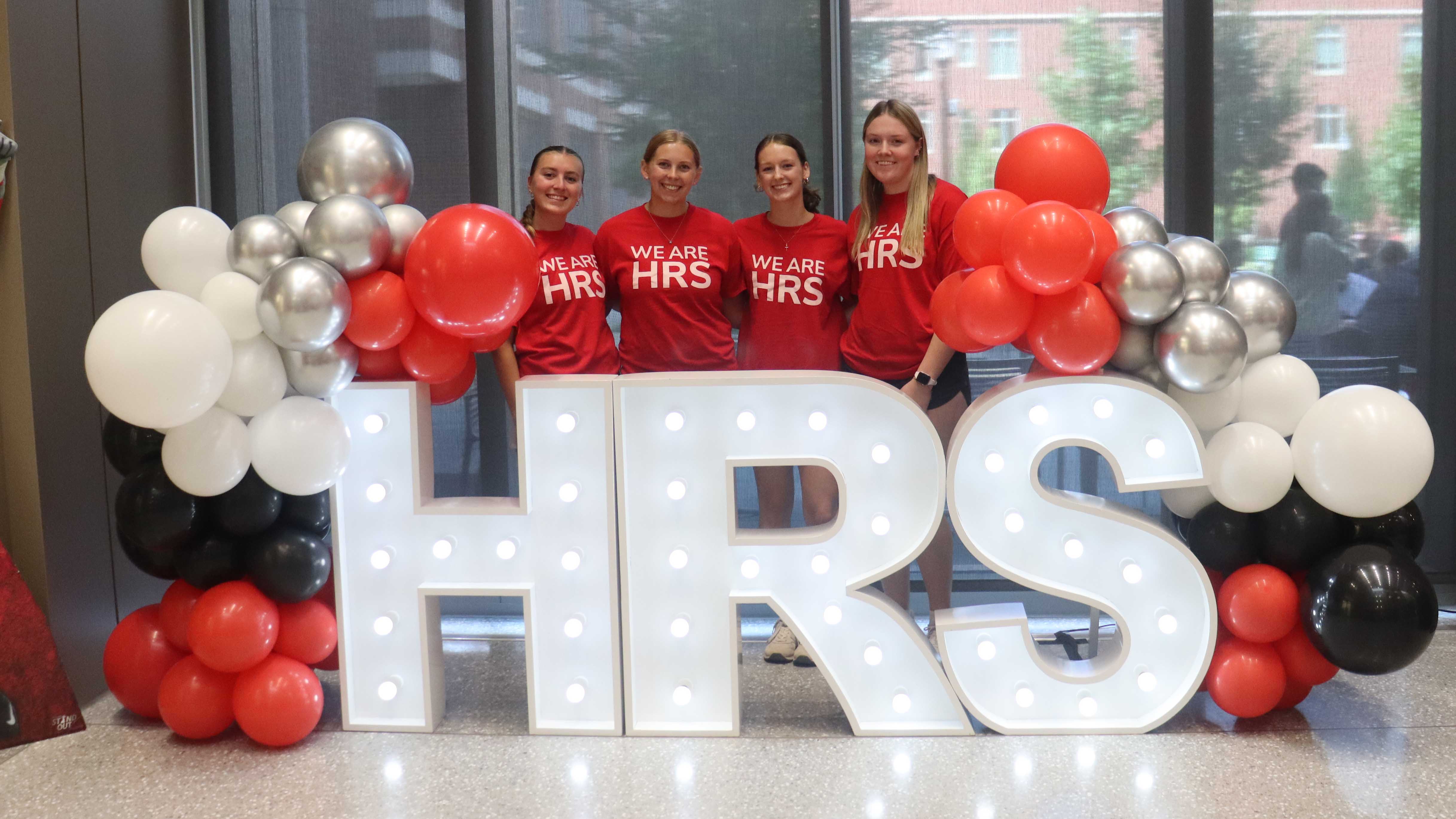 Four students wearing "We are HRS" shirts standing behind decorative letters HRS surrounded by balloons.