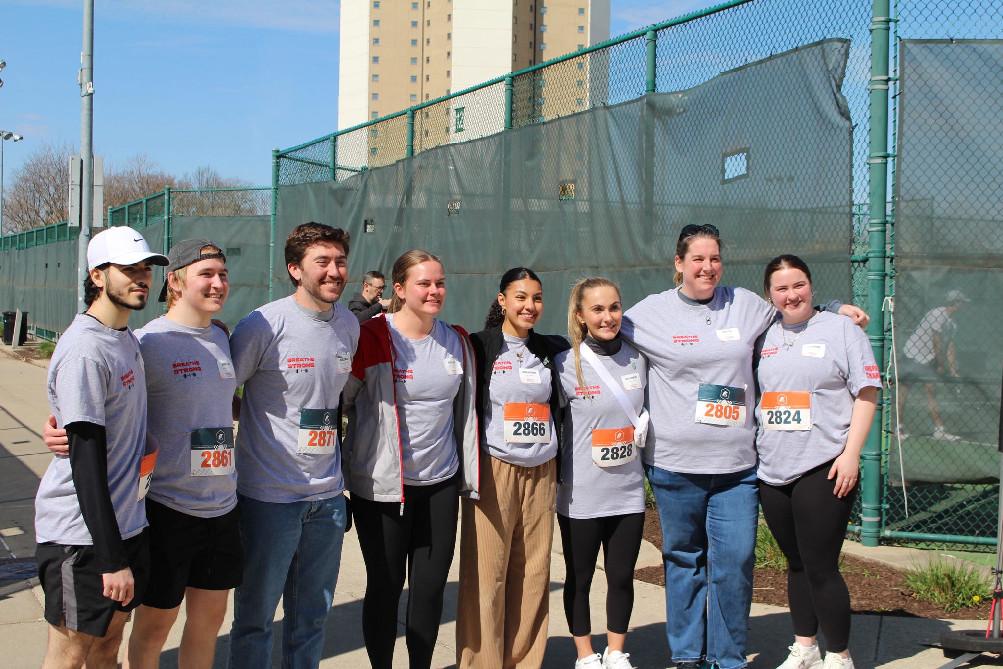 A group of 7 students and 1 faculty member. They are standing together after a walking race for a photo and wearing gray shirts with a logo that reads "Breath Strong".