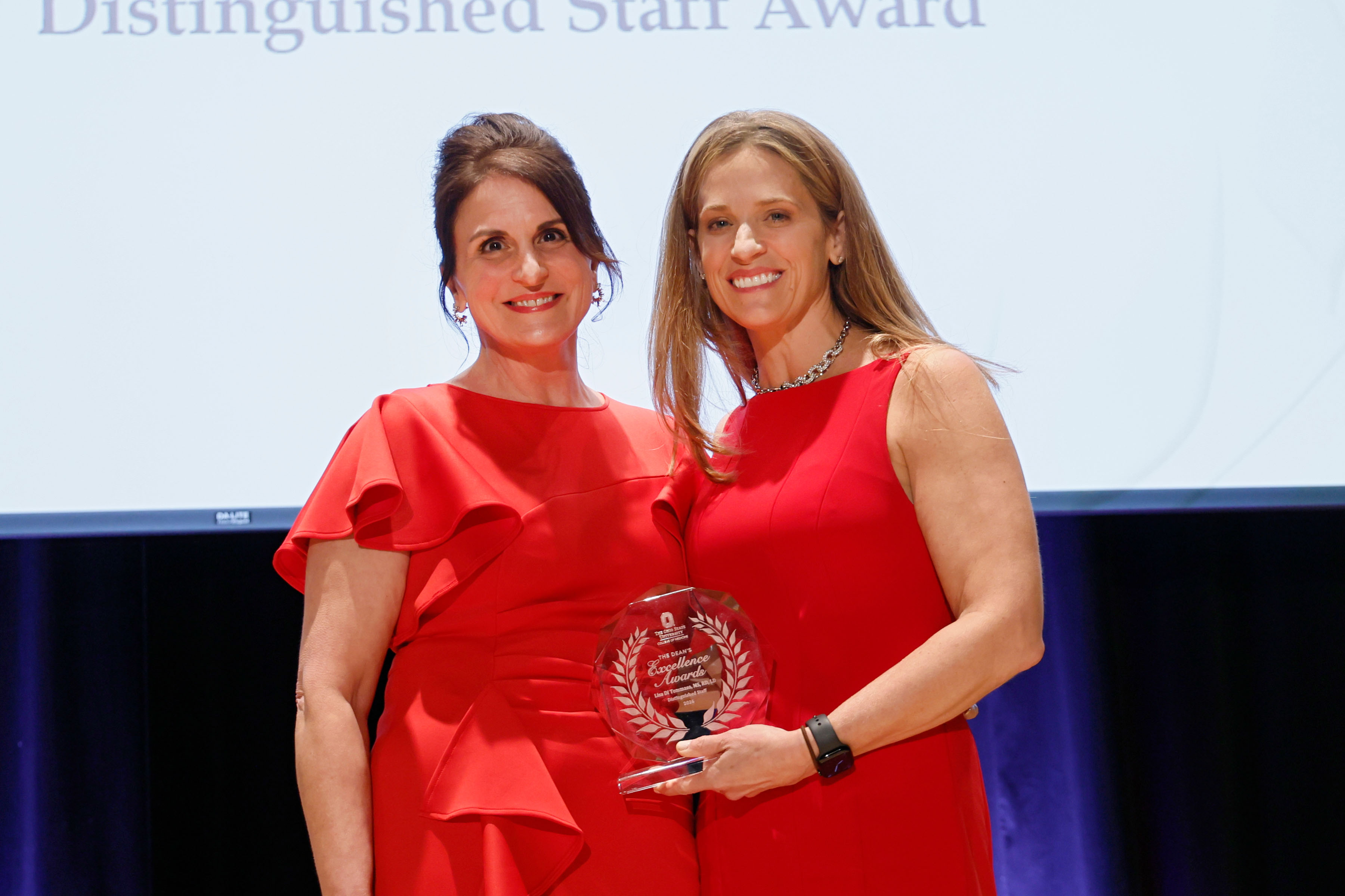 Lisa Di Tommaso standing on the left wearing a red dress and smiling along with Interim Dean Moore, also wearing a red dress and holding an award to be presented to Lisa.