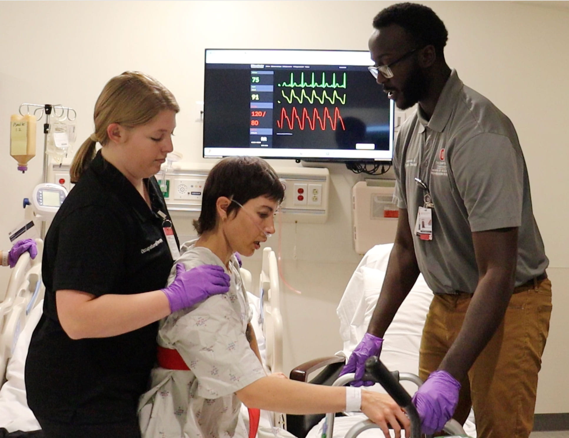 Two medical professionals assist a patient seated on a hospital bed to a standing position using a walker.