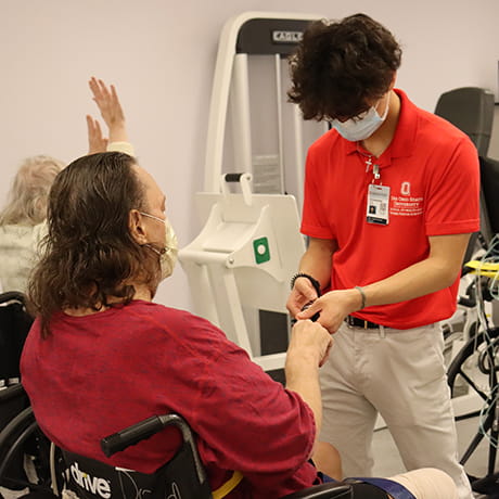 Student in a red polo taking a patient's pulse who is seated in a wheelchair