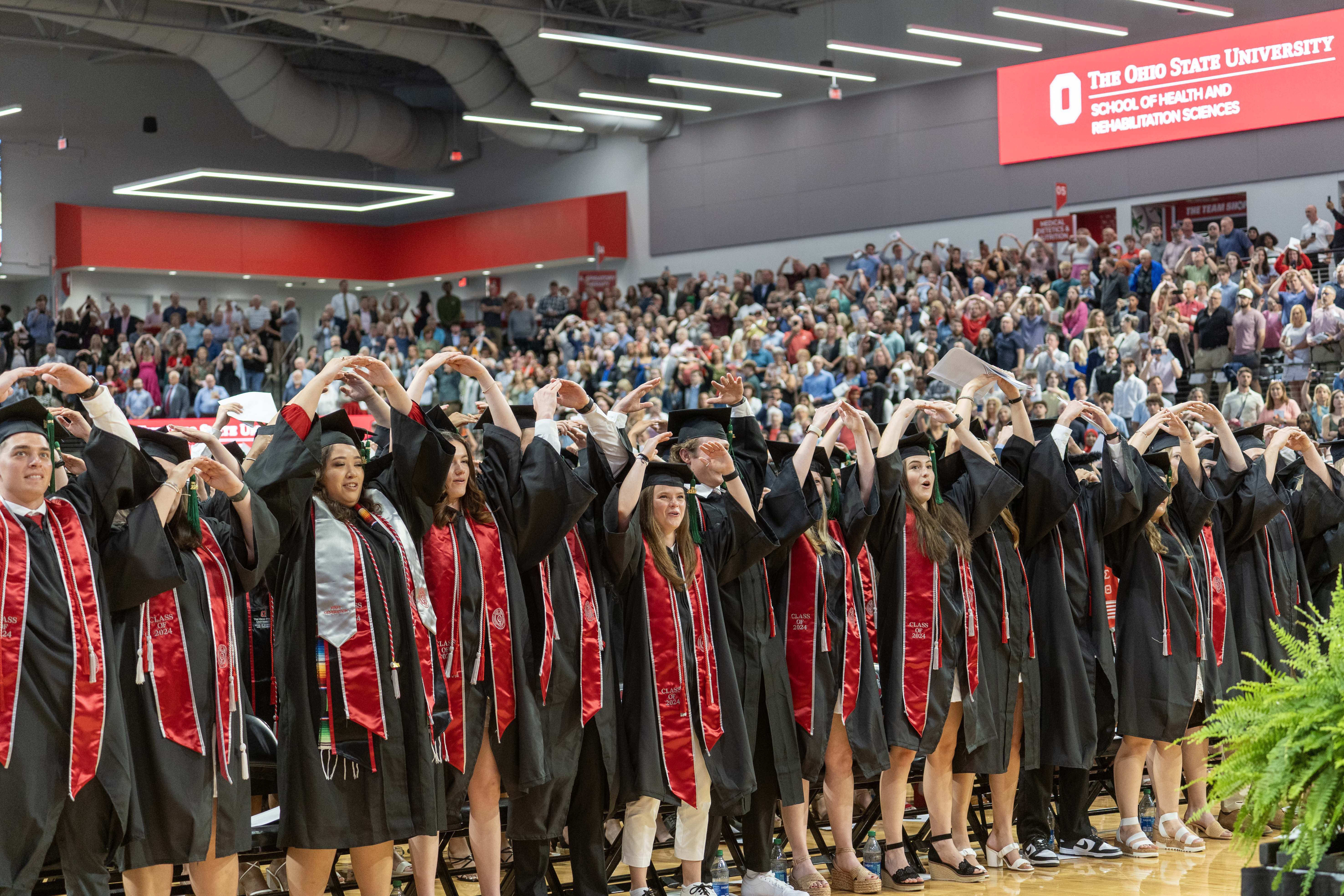 A large group of students wearing graduation caps and gowns, with the hands raised forming an O for Ohio. The students are in the Covelli center auditorium with family and friends seated in bleachers behind them.