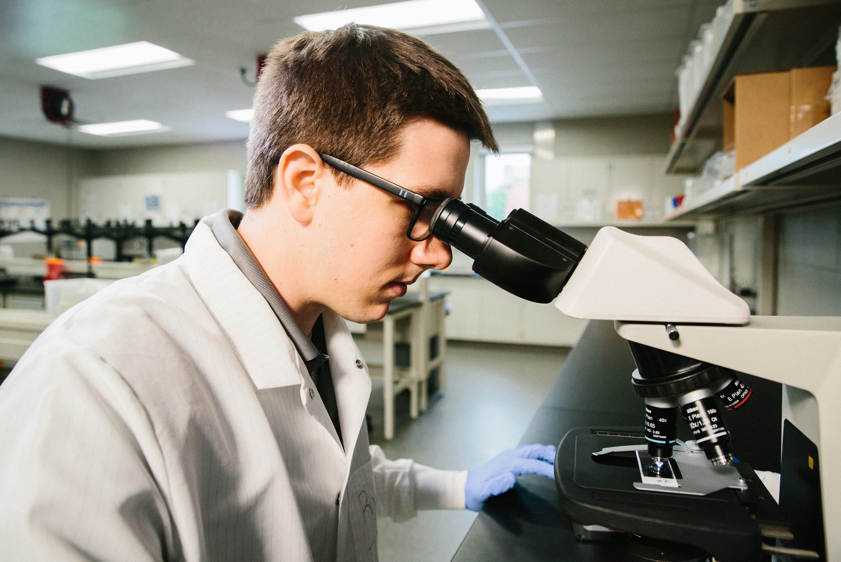 Student in white lab coat looking through a microscope