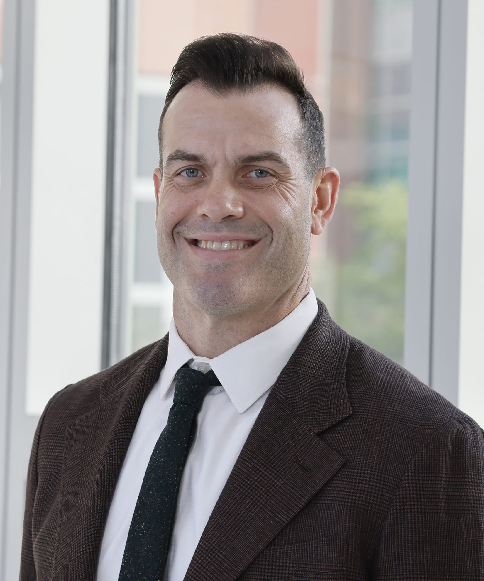 Josh Leonardis wearing a dark suit jacket standing in front of large windows with natural light