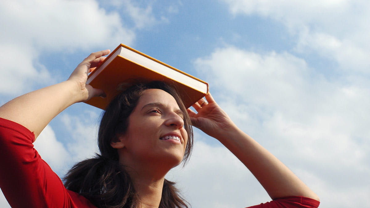 woman balancing book on her head with blue sky and white clouds in background